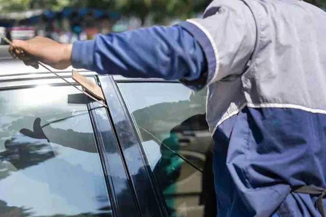A worker in a blue jacket wedges the top of a car door open and reaches inside with a long thin rod.