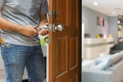A pair of hands turns a key in the lock of an open wooden front door.