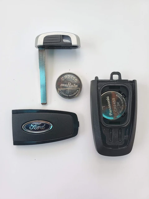 A Ford smart key fob taken apart on a white table. The metal emergency key, the empty fob cover, the open case with a CR2450 coin cell seated inside, and a spare CR2450 cell beside it.