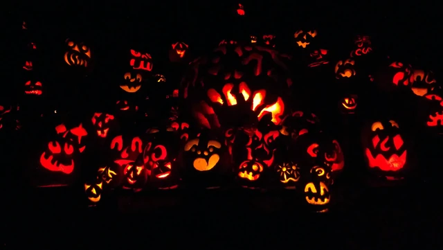 Dozens of carved jack-o-lanterns glowing orange and red in the dark, stacked in rows.